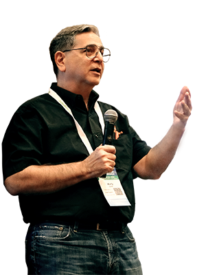 Marty Greif speaking in front of a crowd. Marty has short hair parted to the side, and is wearing glasses. He is dressed in black jeans and a black SiteTuners shirt. Marty is talking with his left hands stretched out with his palm up.  His right hand is holding a microphone in front of his chest.