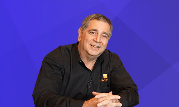 Marty Greif is sitting at a table with his arms resting on it. He is resting his hands in front of him with his fingers interlaced. Marty is smiling while looking at the camera. Marty has short hair parted to the side. He is dressed in a black SiteTuners shirt.